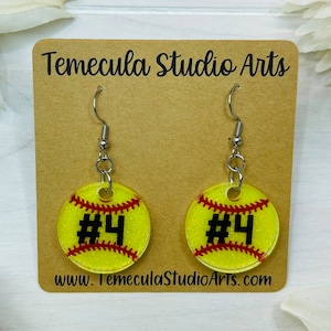 May include: A pair of silver dangle earrings with yellow and red glitter softball charms. The charms have the number "#4" printed in black.
