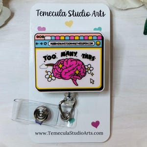 May include: A white badge reel with a cartoon design of a computer window with the text "TOO MANY TABS" and a pink brain. The badge reel has a clear plastic strap and a metal clip. The background is white with small hearts and flowers.