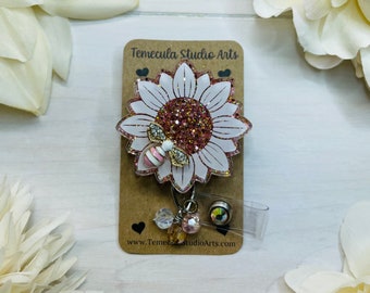 Puede incluir: Un girasol de purpurina rosa y blanco con un dije de abeja dorada en una tarjeta marrón con el texto "Temecula Studio Arts" y "www.TemeculaStudioArts.com". El girasol está unido a un portagafete retráctil transparente con un dije de cristal.