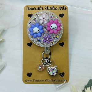 May include: A clear badge reel with a sparkly background and three colourful flowers with smiling faces. The flowers are purple, pink, and white. The reel is attached to a silver chain with a clear plastic badge holder. The text "Temecula Studio Arts" and "www.TemeculaStudioArts.com" are printed on the card.