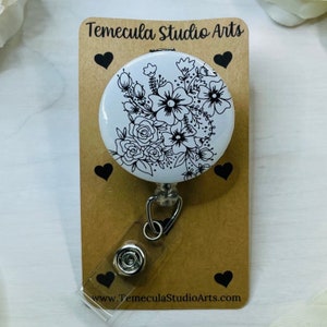 May include: White retractable badge holder with a black and white floral design. The design features a variety of flowers and leaves. The text "Temecula Studio Arts" is printed on the card.