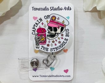 May include: A circular badge reel featuring a skull wearing a pink captain's hat and holding a pink coffee cup. The text reads CAPTAIN OF THE STRUGGLE BUS, surrounded by colourful star icons. The badge is mounted on a white card with Temecula Studio Arts branding.