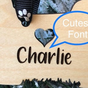Op de afbeelding: Houten kerstversiering in de vorm van een hond met een hartvormige uitsparing en de naam "Charlie" gegraveerd in een schattig lettertype.