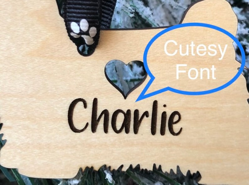 Op de afbeelding: Houten kerstversiering in de vorm van een hond met een hartvormige uitsparing en de naam "Charlie" gegraveerd in een schattig lettertype.