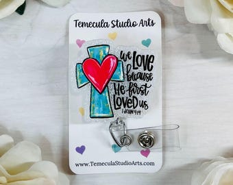 May include: A white card with a glitter background featuring a blue cross with a red heart on top. The text "We love because He first loved us 1 John 4:19" is written in black. The card is attached to a clear badge reel with a silver clip.