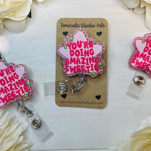 May include: Pink star-shaped badge holder with a glitter finish and the text "You're doing amazing sweetie".