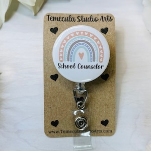 May include: A white round badge with a rainbow design and the text "School Counselor". The badge is attached to a retractable badge holder with a silver clip.