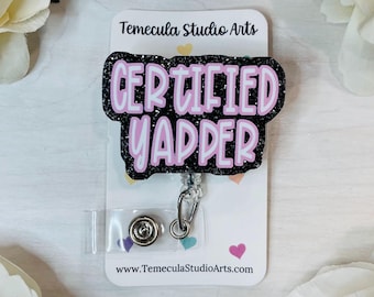 Op de afbeelding: Zwarte glitter badgehouder met een roze "CERTIFIED YAPPER" tekst overlay. De badgehouder is bevestigd aan een transparante intrekbare badgehaspel.