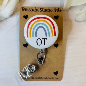 May include: A circular badge reel featuring a white background with a colorful rainbow graphic and the black text OT. The badge is mounted on a rectangular brown card printed with the text Temecula Studio Arts and small black heart icons.