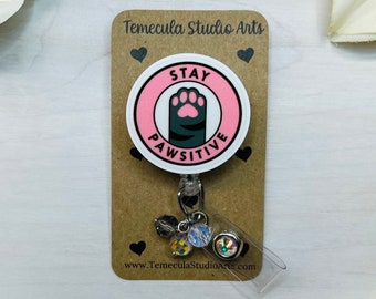 May include: A white and pink round badge with a black paw print and the text "Stay Pawsitive". The badge is attached to a retractable badge holder with a silver clip and a charm with iridescent beads.