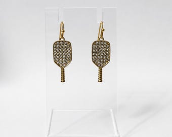 May include: A pair of gold-toned dangle earrings shaped like pickleball paddles. Each paddle features a textured handle and a face encrusted with numerous small, clear, sparkling rhinestones. The earrings hang from simple gold-toned wire hooks.