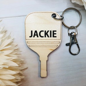 May include: A wooden keychain shaped like a paddle with the name "JACKIE" engraved on it. The keychain has a silver metal ring and a silver metal clasp.
