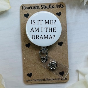 May include: White retractable badge holder with a round white button that says "Is it me? Am I the drama?" The button is attached to a brown card with black hearts and the text "Temecula Studio Arts" and "www.TemeculaStudioArts.com"