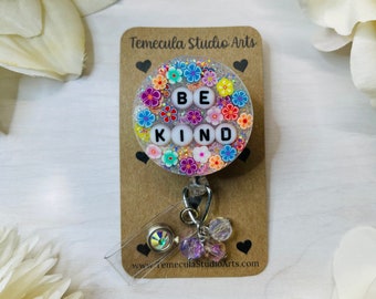 May include: A white and silver badge holder with a clear, round, resin charm that says "BE KIND" in white letters surrounded by colourful flowers. The charm is attached to a silver retractable badge reel with a silver and iridescent crystal charm.