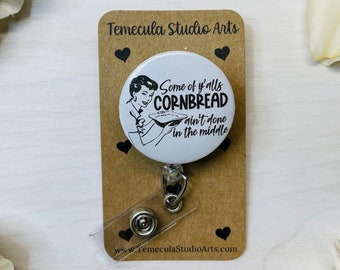 May include: A white badge reel featuring a black and white illustration of a woman with the text "Some of y'alls cornbread ain't done in the middle." The reel is on a brown card with black heart designs.
