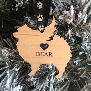 Op de afbeelding: Houten kerstversiering in de vorm van een dwergspits met de naam "BEAR" en een hartvormige uitsparing. De versiering hangt aan een zwart lint met witte poot afdrukken.