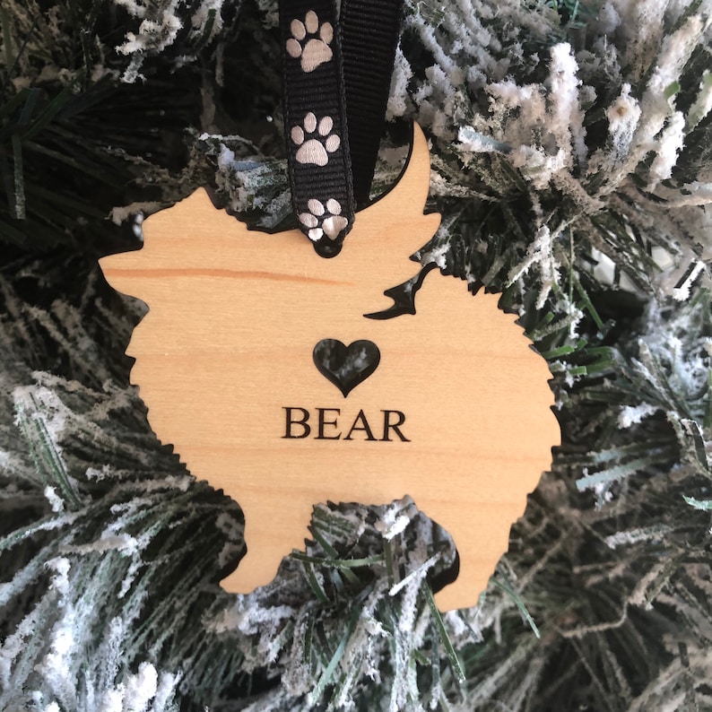 Op de afbeelding: Houten kerstversiering in de vorm van een dwergspits met de naam "BEAR" en een hartvormige uitsparing. De versiering hangt aan een zwart lint met witte poot afdrukken.