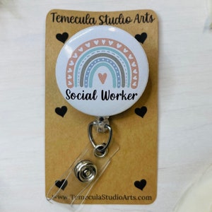 May include: A white Social Worker badge reel with a rainbow design in pastel colors. The badge reel is attached to a brown card with black heart accents and the text "Temecula Studio Arts" and "Social Worker".