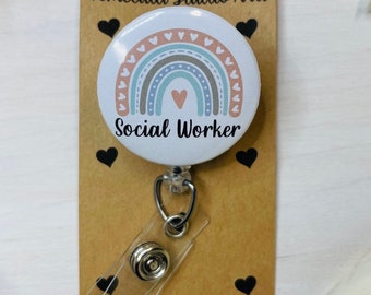 Könnte beinhalten: Ein weißes Social Worker-Ausweis-Rollband mit einem Regenbogen-Design in Pastellfarben. Das Ausweis-Rollband ist an einer braunen Karte mit schwarzen Herzakzenten und dem Text "Temecula Studio Arts" und "Social Worker" befestigt.