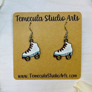 May include: A pair of silver roller skate earrings with white skates, red laces, and blue wheels. The earrings are on a brown card with the text "Temecula Studio Arts" and "www.TemeculaStudioArts.com".