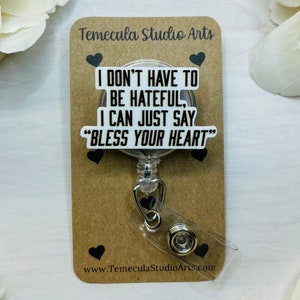 May include: A white badge holder with a black retractable badge reel. The badge holder has the text "I don't have to be hateful, I can just say "Bless your heart"" printed on it. The badge holder is on a brown card with the text "Temecula Studio Arts" and "www.TemeculaStudioArts.com" printed on it.