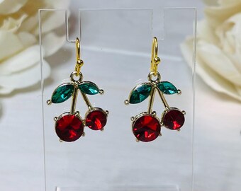 May include: A pair of gold-toned cherry earrings. Each earring features two red, gem-like cherries with green leaves. The earrings are displayed on a clear stand, with a soft background.