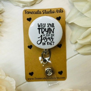 May include: White round badge holder with a black and white graphic that reads "Why y'all tryin' to test the Jesus in me?" The badge holder is on a brown card with the text "Temecula Studio Arts" and a website address.