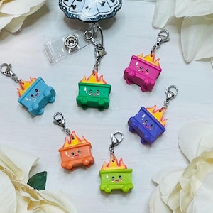 May include: A collection of colorful, miniature dumpster charms, each with a smiling face and flames. The charms are in various colors: blue, green, pink, purple, and orange. Each charm is attached to a silver-colored clasp.