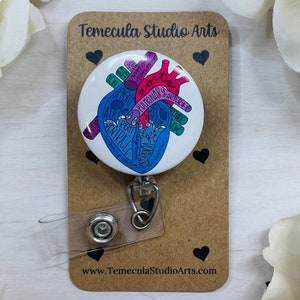 May include: A white heart-shaped badge reel with a colorful anatomical illustration of the human heart. The heart is labeled with anatomical terms such as "aorta", "pulmonary artery", "right ventricle", and "left atrium". The badge reel is attached to a clear plastic strap with a metal clip. The background is a brown card with the text "Temecula Studio Arts" and "www.TemeculaStudioArts.com".