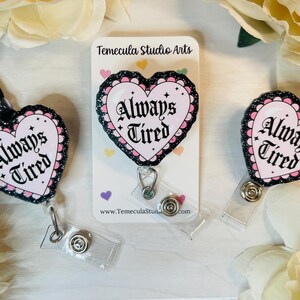 Peut inclure: Trois porte-badges en forme de c&oelig;ur noir et rose avec le texte "Always Tired" en lettres noires. Les porte-badges sont sur un fond blanc avec une carte de visite qui dit "Temecula Studio Arts".