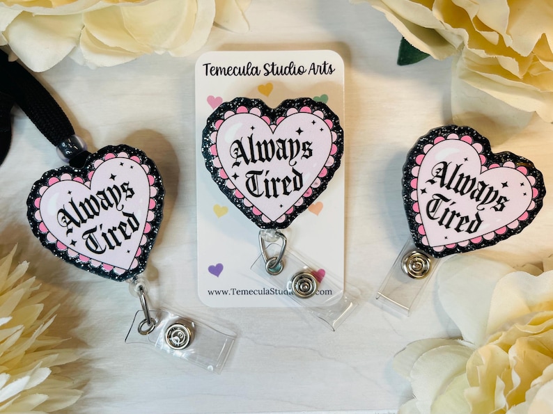 Peut inclure: Trois porte-badges en forme de c&oelig;ur noir et rose avec le texte "Always Tired" en lettres noires. Les porte-badges sont sur un fond blanc avec une carte de visite qui dit "Temecula Studio Arts".