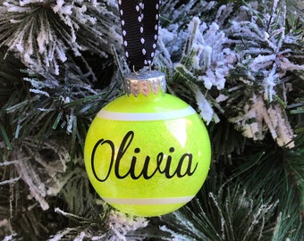 May include: A yellow and white tennis ball Christmas ornament with the name "Olivia" written in black cursive.