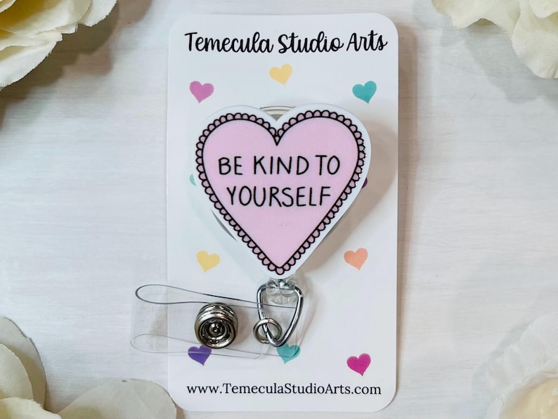 May include: A pink heart-shaped badge reel with the text "BE KIND TO YOURSELF" in black. The badge reel has a decorative border and a retractable cord. The background is white with colorful heart accents and the Temecula Studio Arts logo.