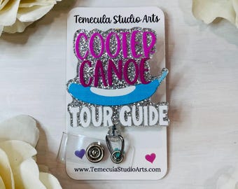 May include: A silver glitter badge reel featuring the text COOTER CANOE in pink and TOUR GUIDE in white, with a light blue canoe graphic in the centre. The badge is mounted on a white display card labelled Temecula Studio Arts at the top.