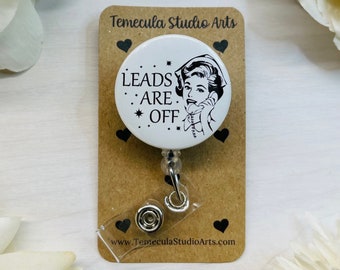 May include: A white circular badge reel featuring a black graphic of a person talking on a telephone with the text LEADS ARE OFF. The badge is attached to a rectangular brown card printed with Temecula Studio Arts and small black hearts.