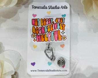 May include: A retractable badge reel with the text "NOT BOSSY, JUST AGGRESSIVELY HELPFUL" in colorful bubble letters. The reel is white with colorful hearts and flowers. The website www.TemeculaStudioArts.com is printed at the bottom.