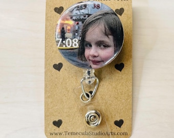 May include: A circular badge reel featuring a photographic print of a young person with a side-glancing expression, overlaid with the white text 7:08. The badge is mounted on a tan card labelled Temecula Studio Arts with small black heart icons.