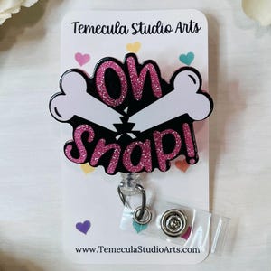 May include: A badge reel with the words "Oh Snap!" in pink glitter, with a white bone graphic. The badge is black with a white border and is attached to a clear plastic reel with a metal clip. The text "Temecula Studio Arts" is at the top.