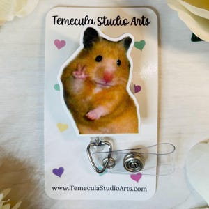 May include: A retractable badge reel featuring a hamster with a peace sign. The hamster is brown and white, with a pink nose. The badge reel is attached to a white card with the text "Temecula Studio Arts" and colorful hearts.