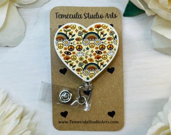 May include: A white heart-shaped badge holder with a colorful retro pattern featuring peace signs, rainbows, eyes, and flowers. The badge holder is attached to a retractable badge reel. The text "Temecula Studio Arts" and "www.TemeculaStudioArts.com" are printed on the card.