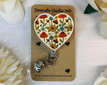 May include: A white heart-shaped badge holder with a colorful psychedelic mushroom and flower pattern. The badge holder is attached to a clear retractable badge reel with a silver clip. The card behind the badge holder says "Temecula Studio Arts" and "www.TemeculaStudioArts.com".