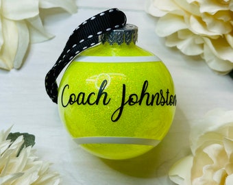 Puede incluir: Un adorno de bola de tenis amarillo y blanco con purpurina. El adorno tiene una cinta negra y el texto "Coach Johnston" escrito en él.