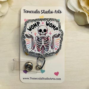 May include: A silver glitter badge reel with a white skeleton design. The skeleton is wearing a pink and blue shirt and has the words "Womp Womp" written above it.