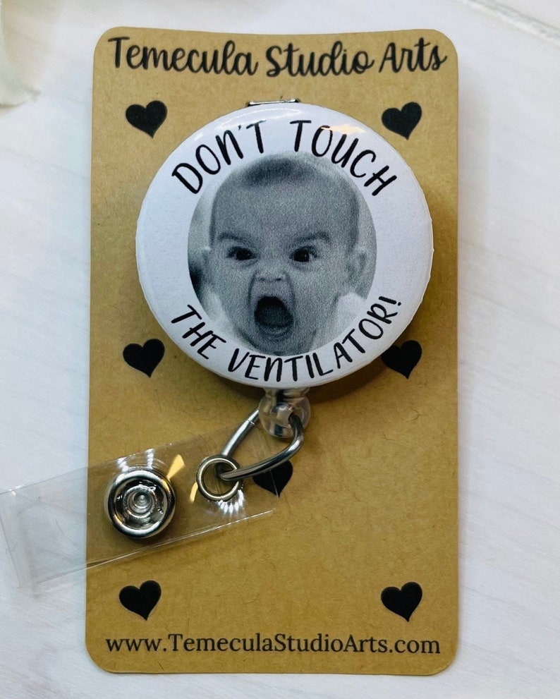 May include: A white button with a black border and the text "Don't Touch The Ventilator!" with a photo of a baby in the center. The button is attached to a retractable badge holder with a clear plastic cover. The background is brown with black hearts.
