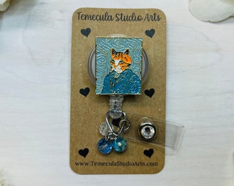 May include: A brown card with black hearts and the text "Temecula Studio Arts" and "www.TemeculaStudioArts.com". A gold retractable badge holder with a blue and orange cat illustration, blue and clear crystal beads, and a clear crystal charm.