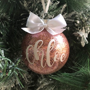 May include: A pink glitter Christmas ornament with a white bow and the name "Chloe" written in white script.