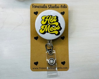 May include: A white badge reel with the words "Hot Mess" in yellow and black lettering. The badge reel is attached to a brown card with black heart designs and the text "Temecula Studio Arts".