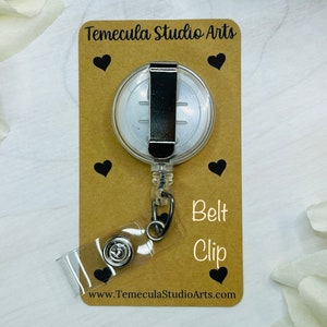 May include: A clear retractable badge holder with a silver belt clip. The holder is on a brown card with the text "Temecula Studio Arts" and "Belt Clip" printed on it. The card also has a website address printed on it: www.TemeculaStudioArts.com.