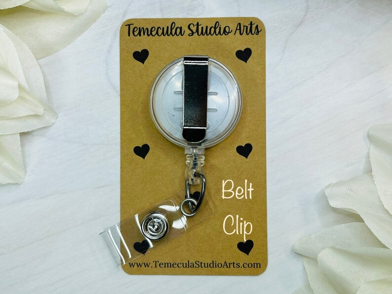 May include: A clear retractable badge holder with a silver belt clip. The holder is on a brown card with the text "Temecula Studio Arts" and "Belt Clip" printed on it. The card also has a website address printed on it: www.TemeculaStudioArts.com.