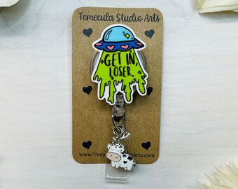 May include: A green, yellow, and blue cartoon UFO with the text "Get in, Loser." on a brown card with black hearts. A white cow charm is attached to a retractable badge holder.